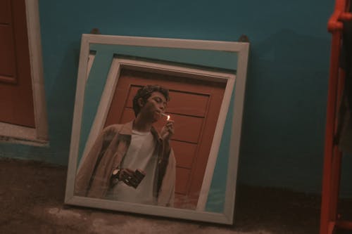 Free Square mirror placed on floor in room and reflecting young rebellious Asian guy in stylish outfit smoking cigarette near door Stock Photo